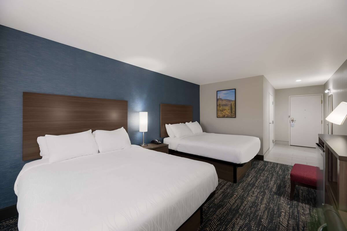 A hotel room with two beds, a desk, a chair, a red bench, a painting on the wall, and a door.