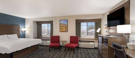 A hotel room with a bed, two red chairs, a desk, a TV, and a painting on the wall.
