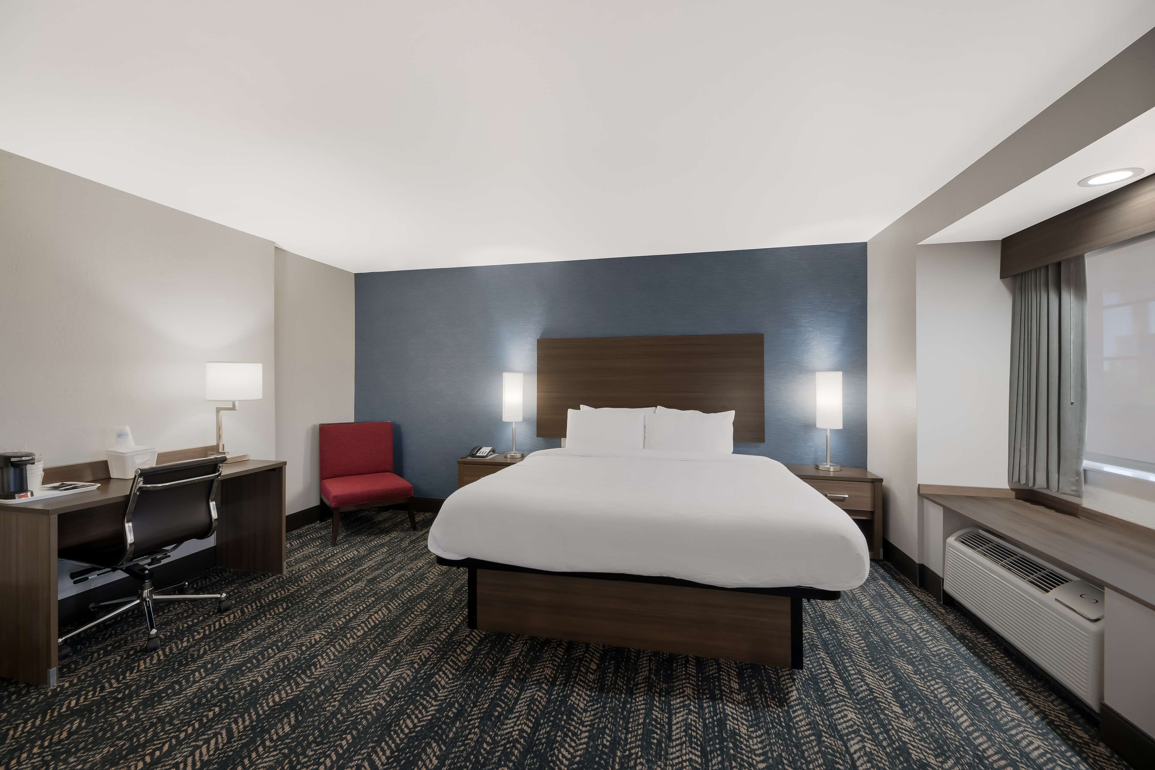 A hotel room with a large bed, a desk with a chair, a red chair, two bedside lamps, and a large window with curtains.