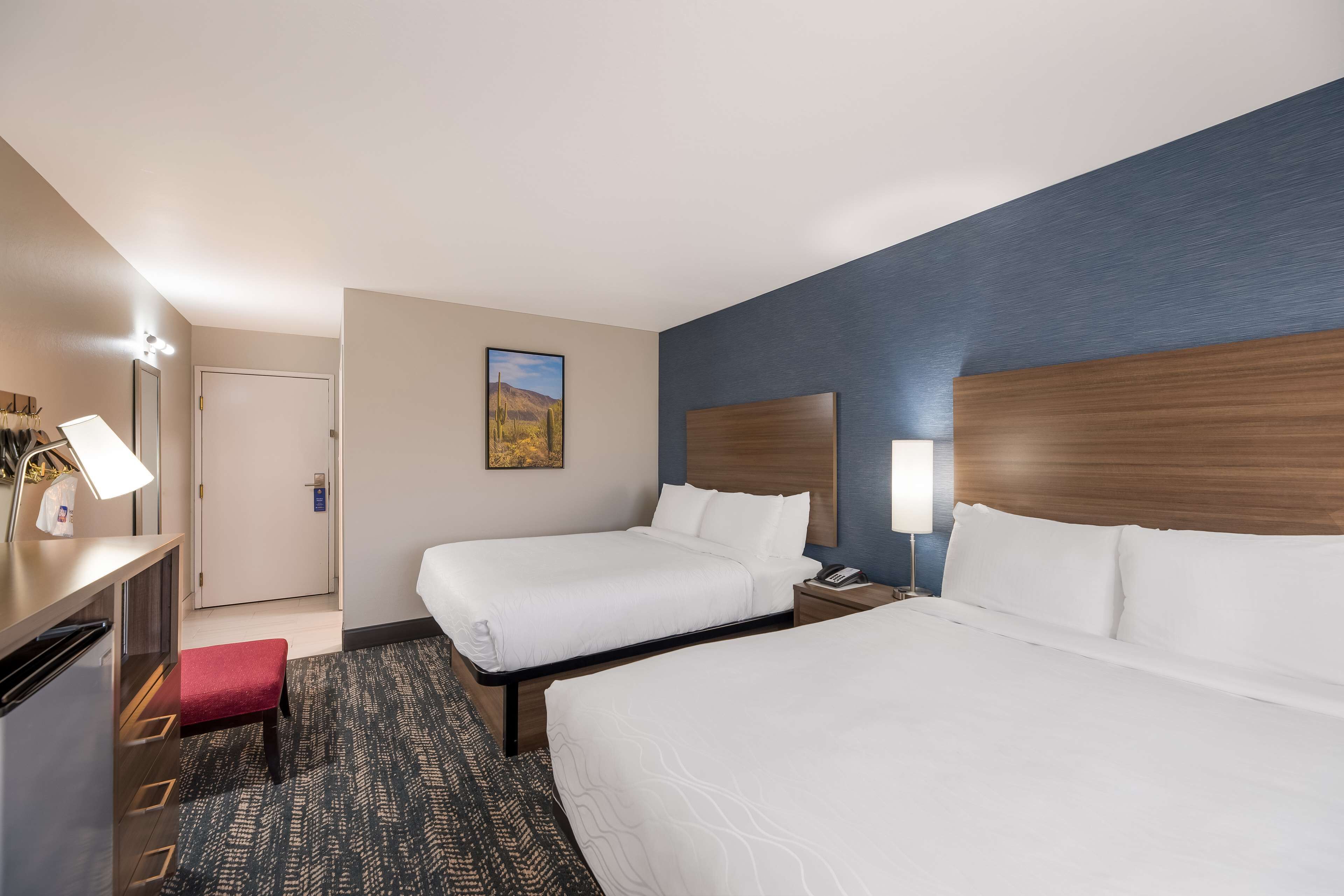 A hotel room with two beds, a desk with a lamp, a red bench, a painting on the wall, and a door.
