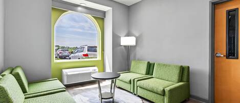 A modern waiting area with green upholstered chairs, a small round table, and a view of parked cars through a large window.