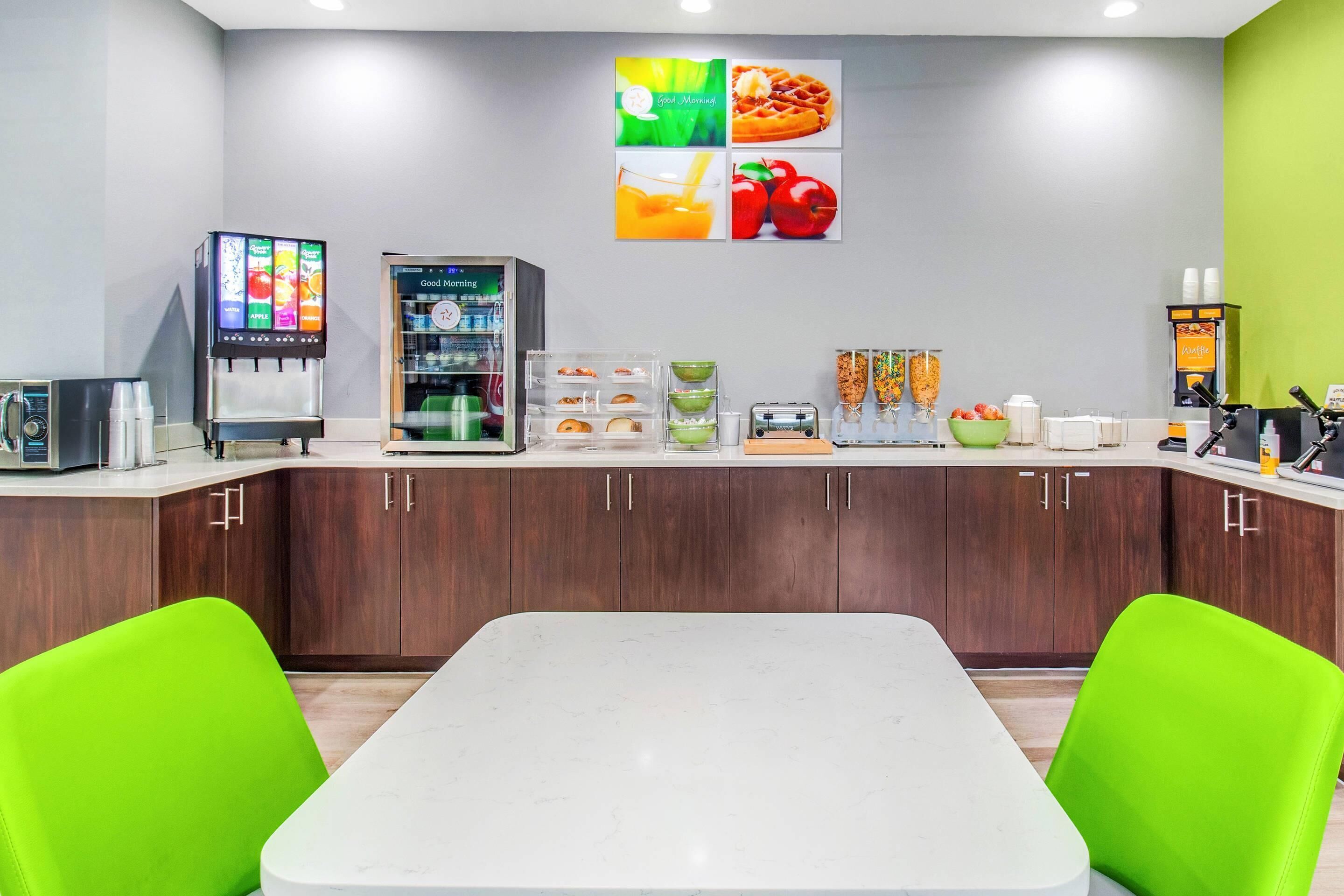 Una cocina moderna con encimera blanca, sillas verdes, cafetera, refrigerador y una pared decorada con ilustraciones de comida.