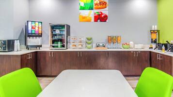 Un espace cuisine moderne avec un plan de travail blanc, des chaises vertes, une machine à café, un réfrigérateur et un mur décoré d’œuvres d’art sur le thème de la nourriture.