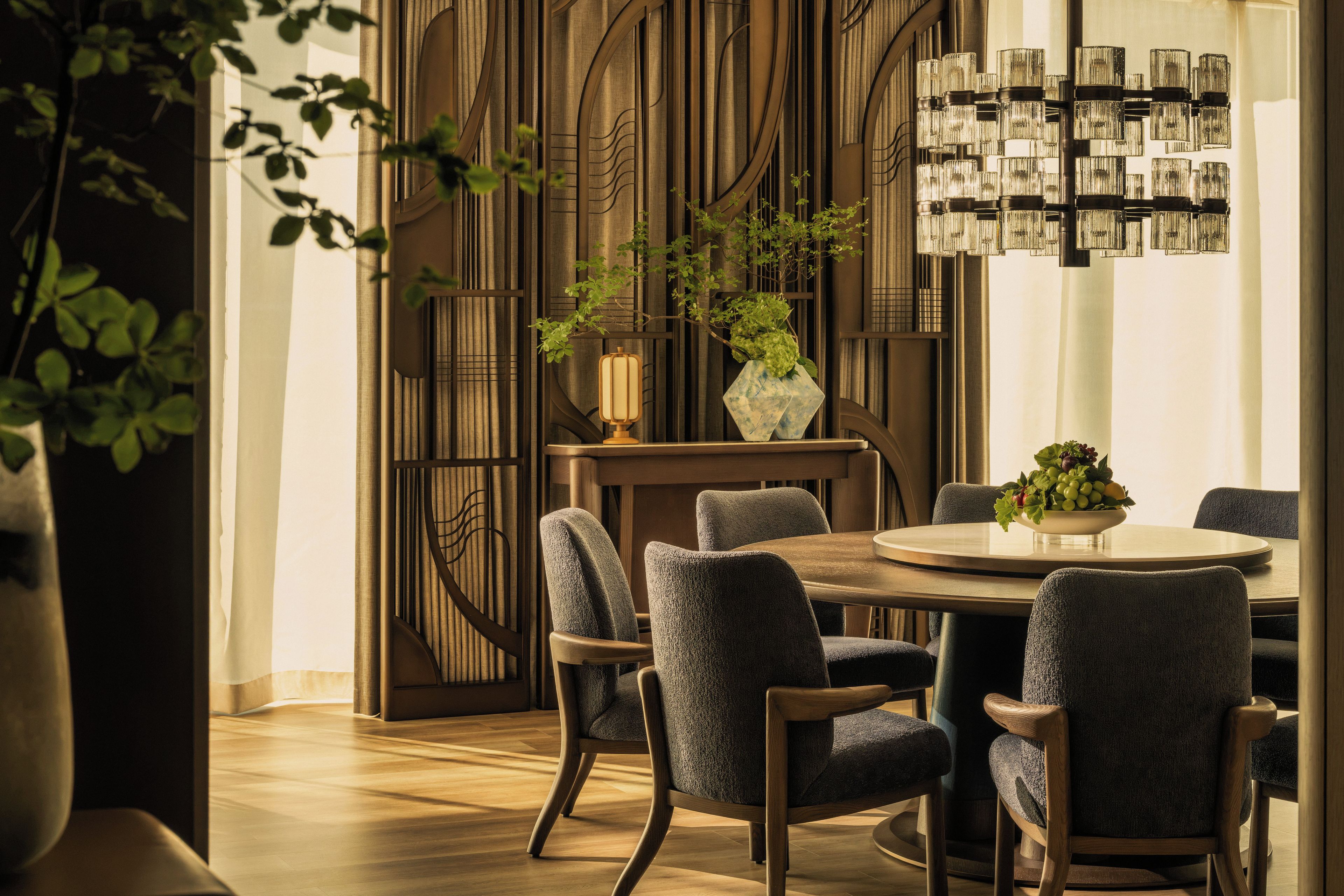 A dining area with a round table, six chairs, a vase with plants, and a wall with vertical lines and a chandelier.