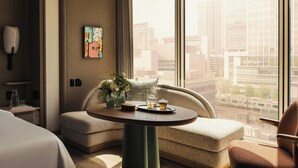 Quarto de hotel moderno com um sofá curvo, uma mesa redonda com bandeja, um vaso com flores e vista para a cidade através de grandes janelas.