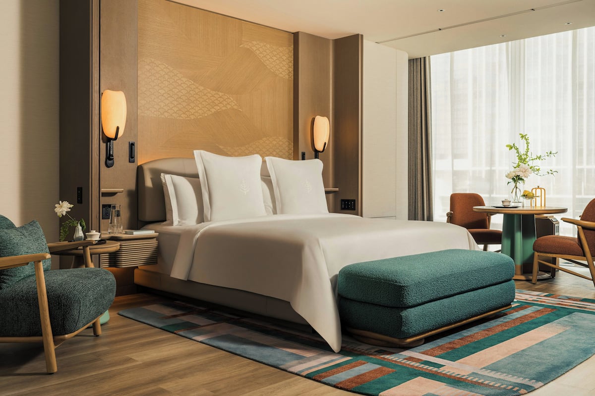 Une chambre d’hôtel moderne dotée d’un grand lit, d’un coin salon avec une table et des chaises, d’une vue sur la ville grâce à de grandes fenêtres et d’un panneau décoratif mural.