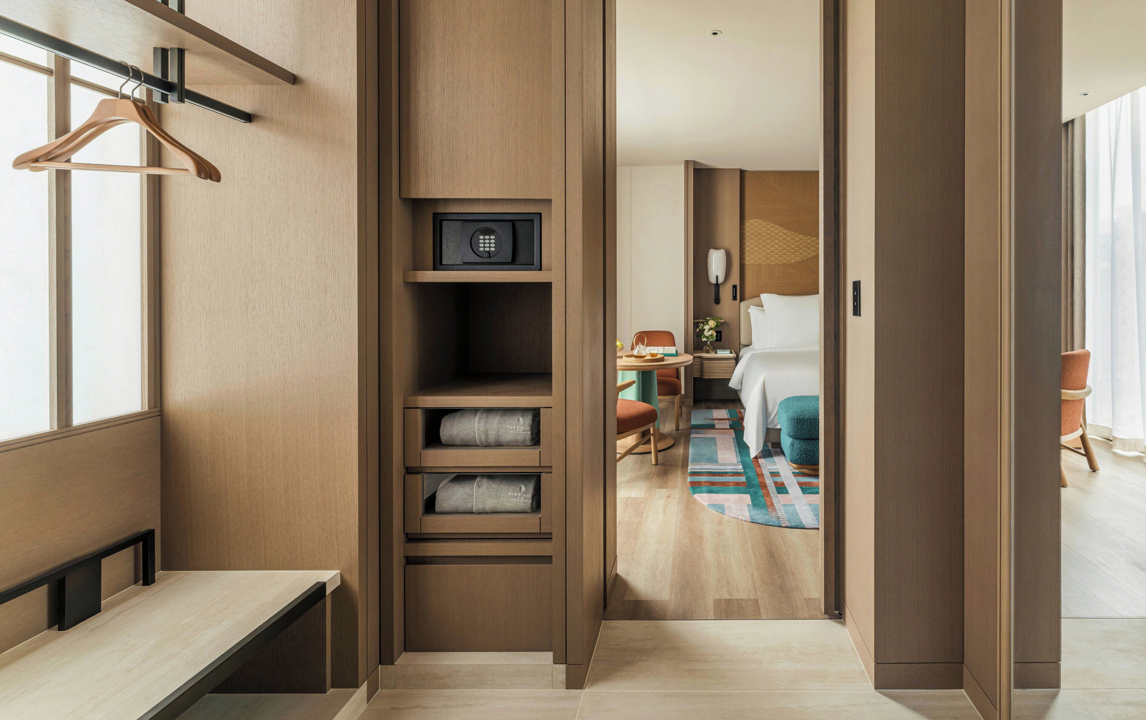 A hotel room with a wooden wardrobe, a bed, a desk, and a bathroom visible through an open door.