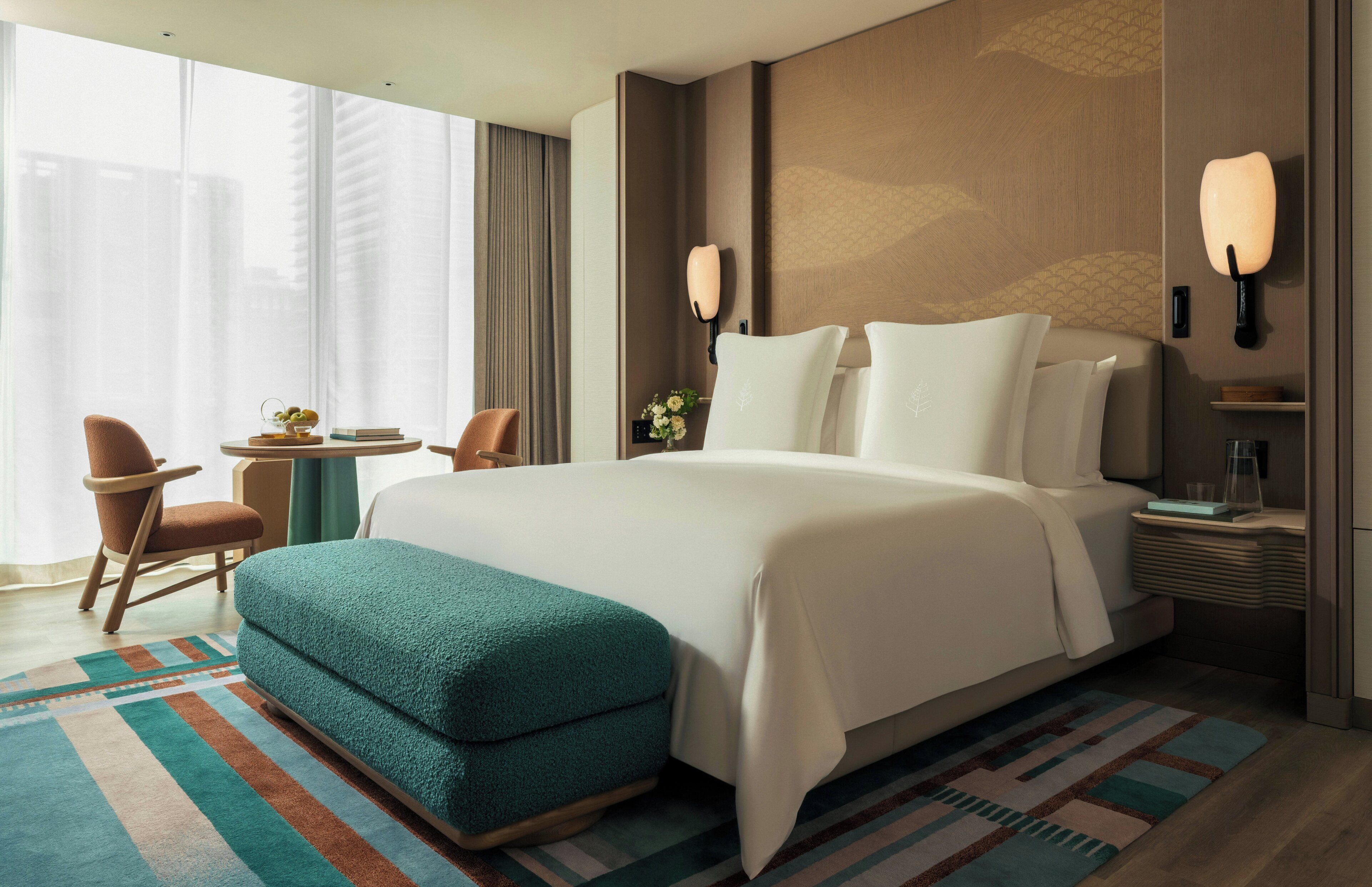 Ein Hotelzimmer mit einem großen Bett, einer Sitzecke mit Sessel und Tisch, einer Nachttischlampe und Blick auf die Stadt.