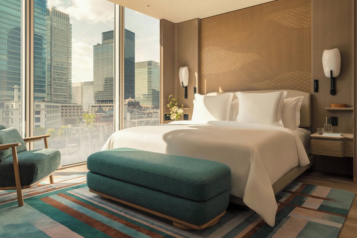 Une chambre d’hôtel moderne avec un grand lit, une vue sur le paysage urbain, un banc recouvert d’un tissu vert et une table de chevet avec une lampe.
