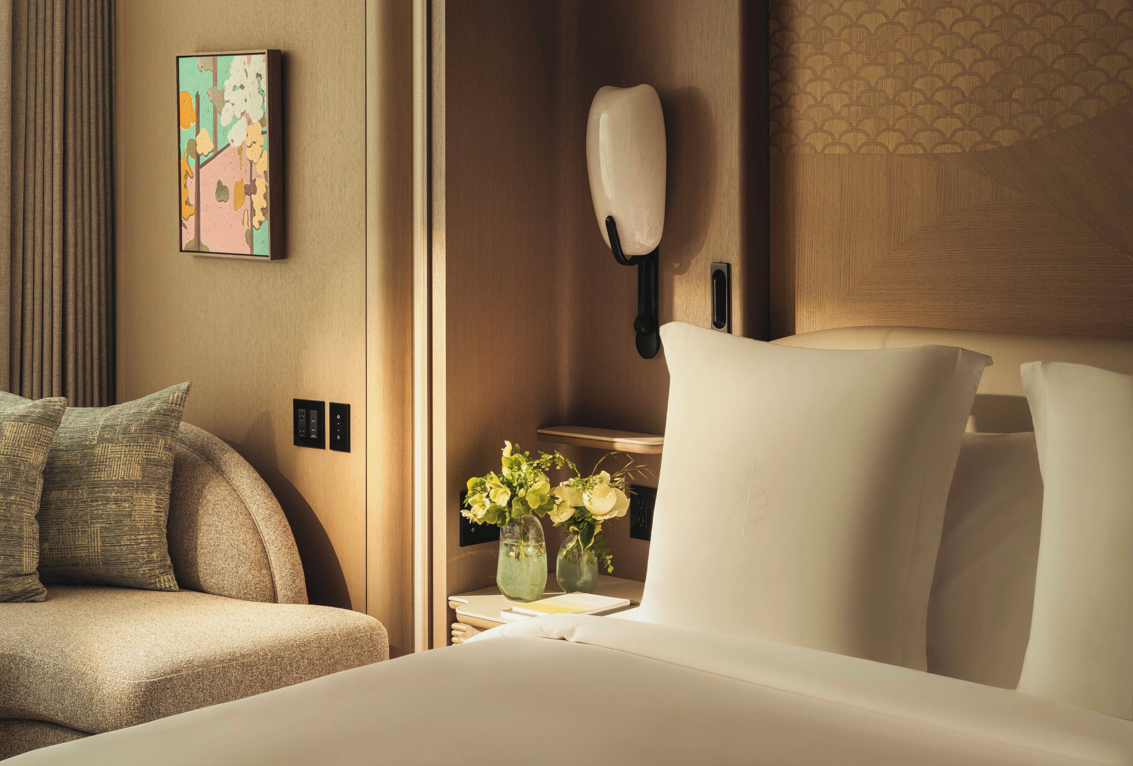 A hotel room with a bed, a nightstand with a vase of flowers, a wall-mounted light, and a painting on the wall.