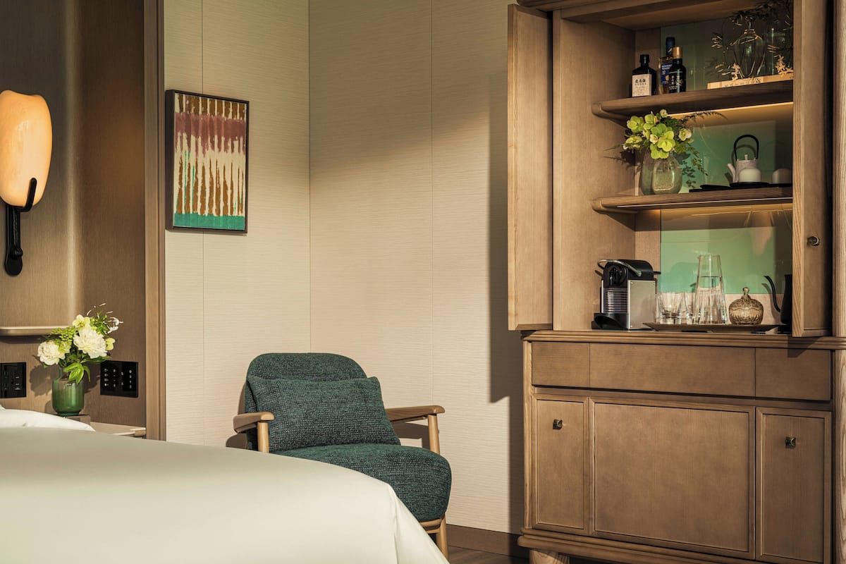 Une chambre d’hôtel comprenant un lit, une chaise, une lampe fixée au mur, un tableau et une armoire en bois avec des étagères.