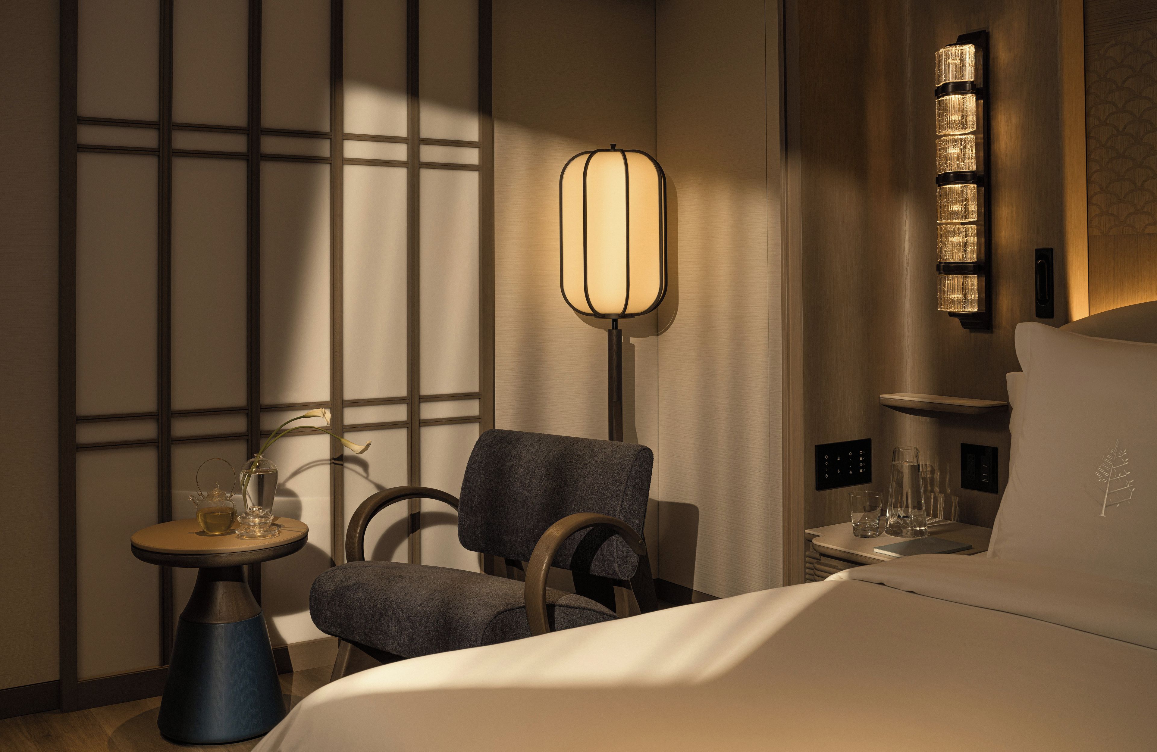 A hotel room with a bed, a chair, a small table with a vase and a teapot, and a wall-mounted lamp.