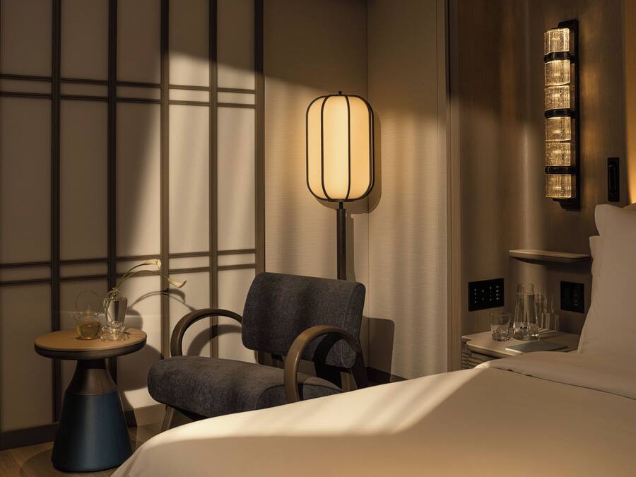 A hotel room with a bed, a chair, a small table with a vase and a teapot, and a wall-mounted lamp.