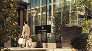 Um homem caminhando em direção à entrada de um hotel, com uma placa indicando 'Four Seasons Hotel Tokyo at Marunouchi'.