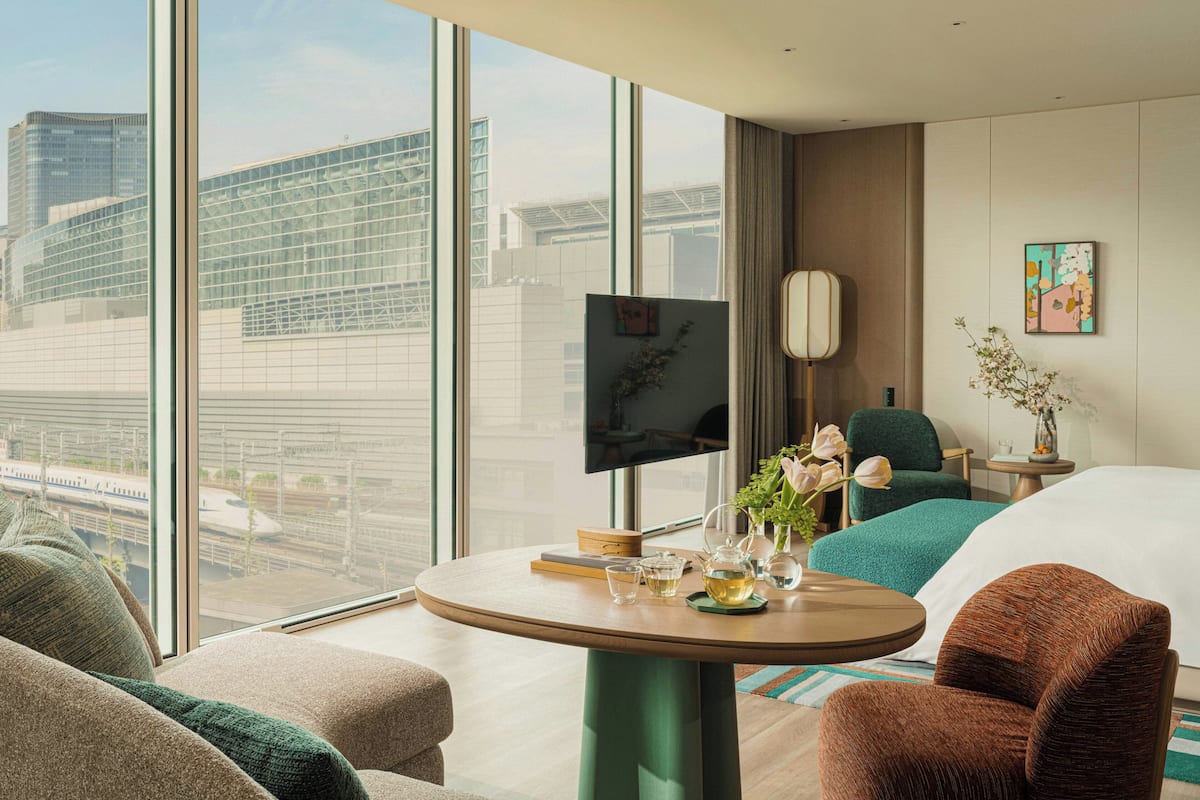 A modern hotel room with a large window, a flat-screen TV, a round table with a vase of flowers, and a comfortable seating area.