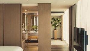 Quarto de hotel moderno com uma cama grande, um espaço de estar, banheiro com toucador e uma janela ampla.