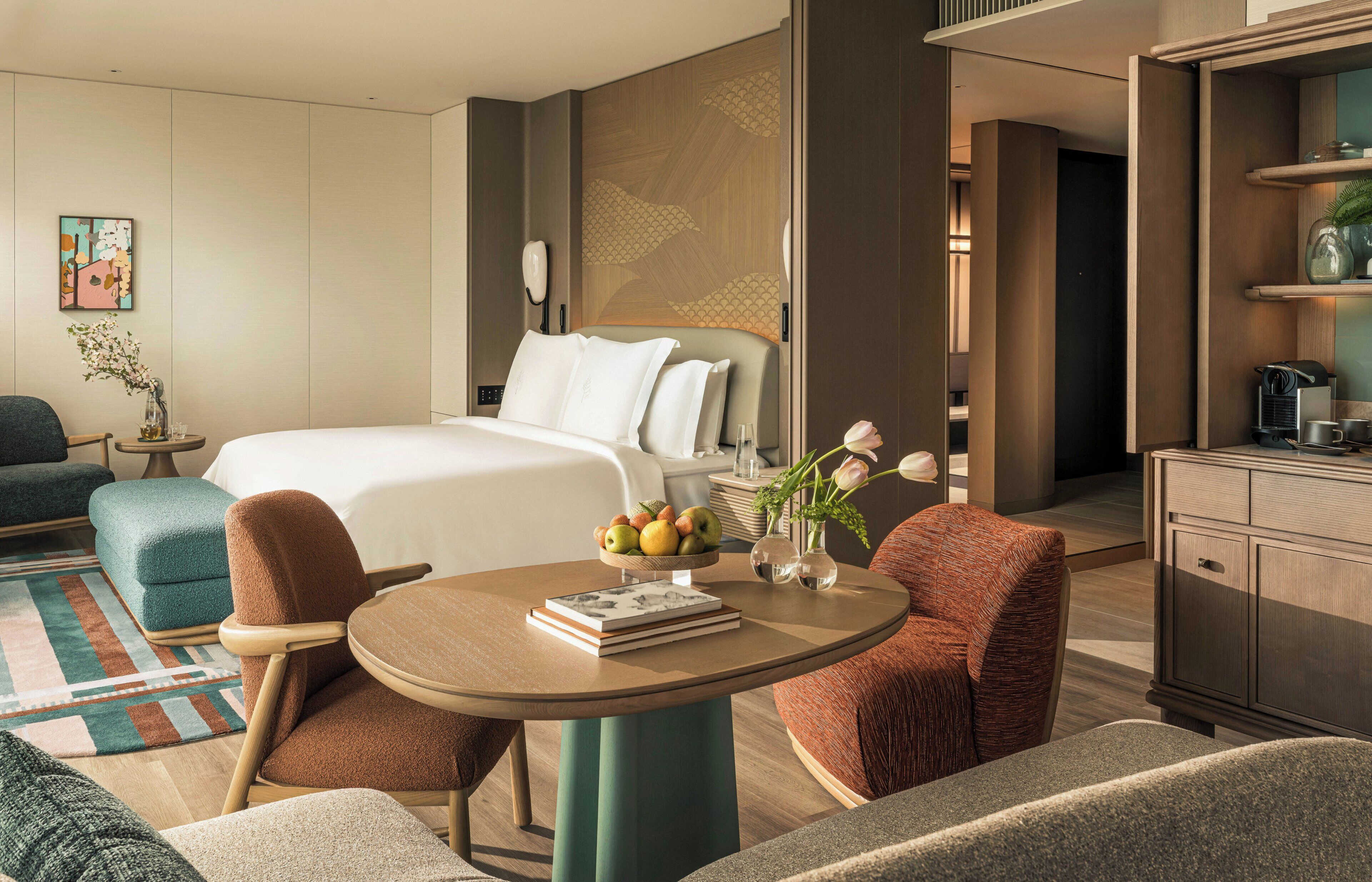 A modern hotel room with a bed, seating area, and a dining table with a fruit bowl.