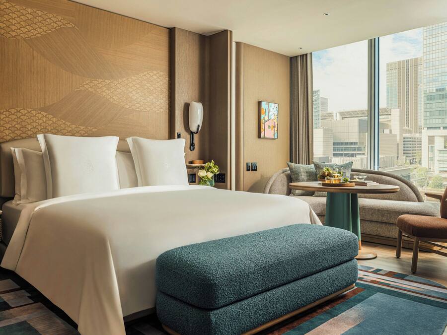 A hotel room with a large bed, a small table, a chair, and a view of the city.