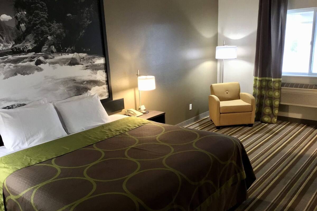 Una habitación de hotel con cama, una silla, un escritorio con lámpara y un cuadro grande en la pared.