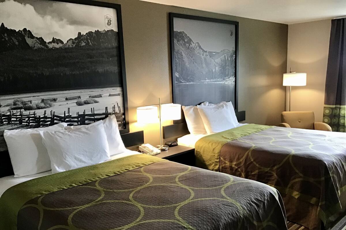 Habitación de hotel con dos camas, una mesita de noche, una lámpara y dos cuadros con paisajes de montañas.