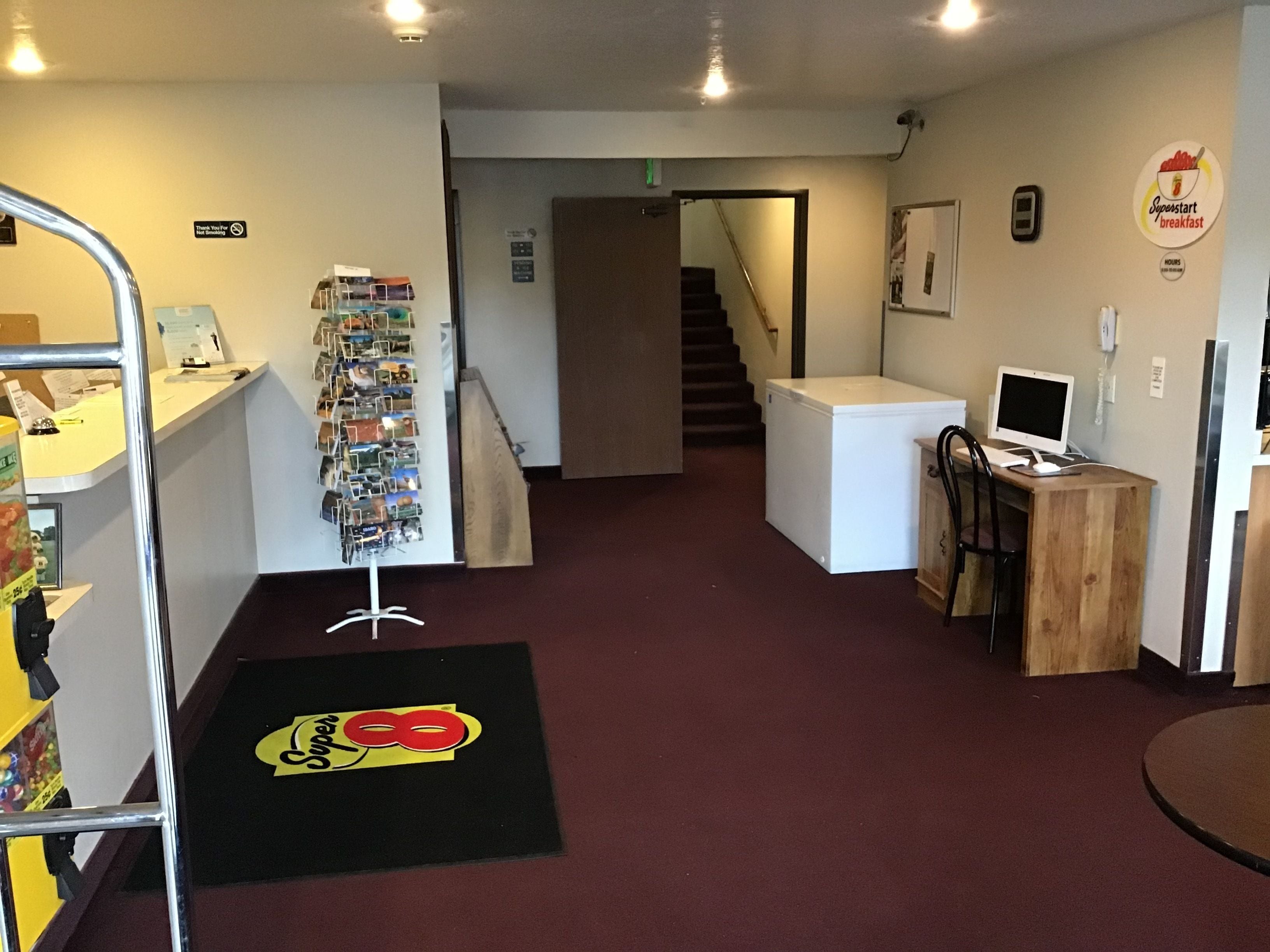 A hotel lobby with a reception desk, a wall-mounted display, a staircase, and a small office area with a computer.