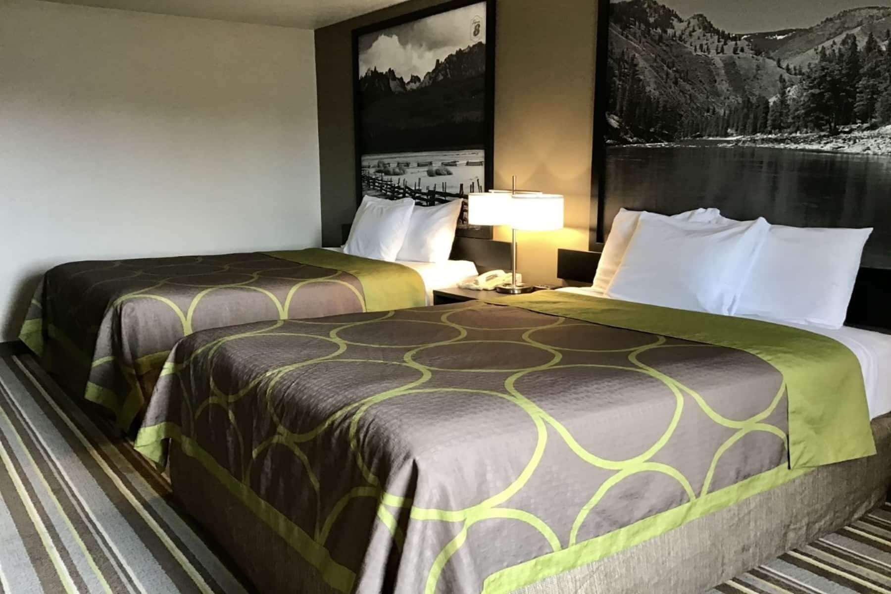 Een hotelkamer met twee bedden, een nachtkastje, een lamp en een groot schilderij van een berglandschap aan de muur.