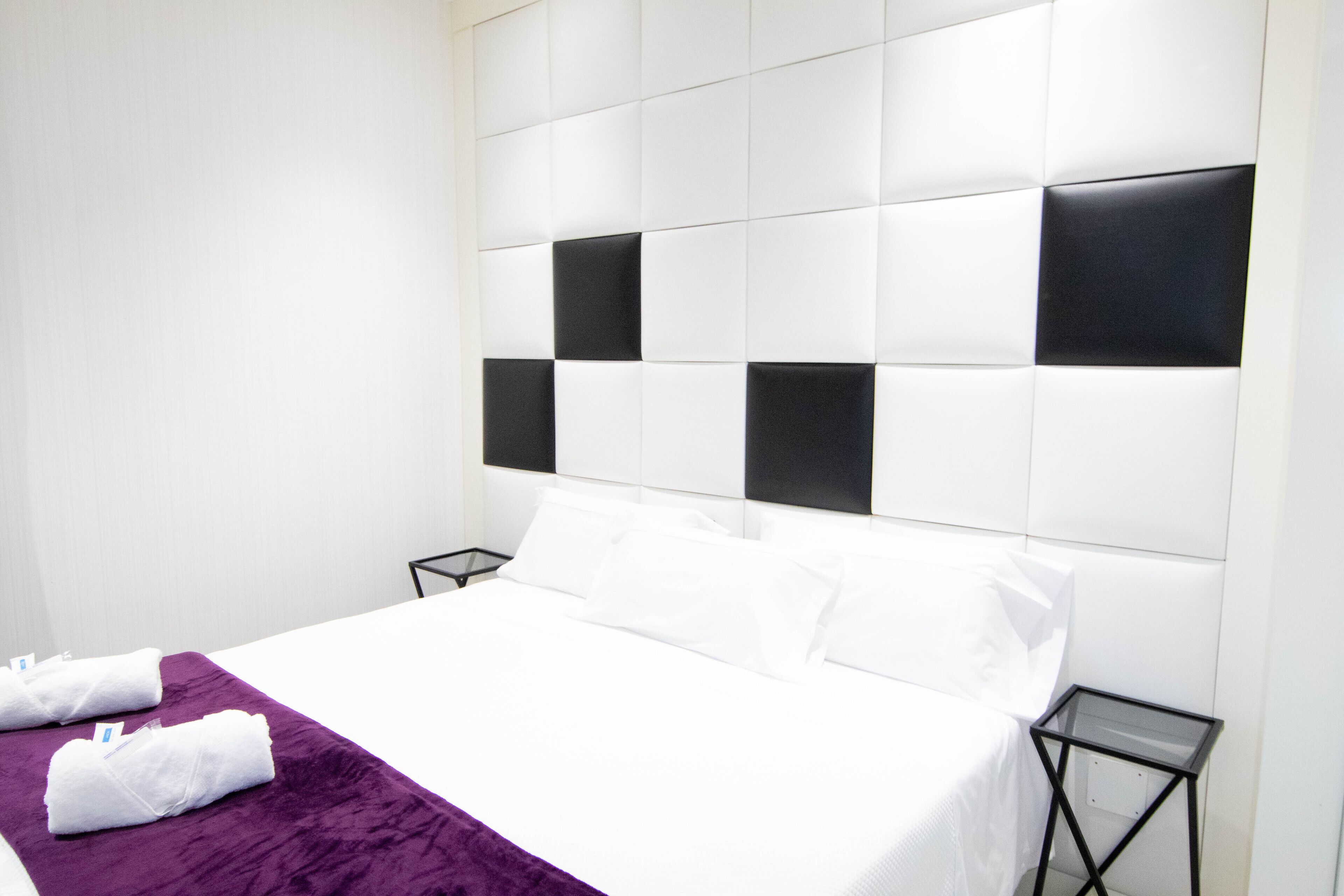 Habitación de hotel con una cama grande, ropa de cama blanca, una manta morada y un cabecero moderno.