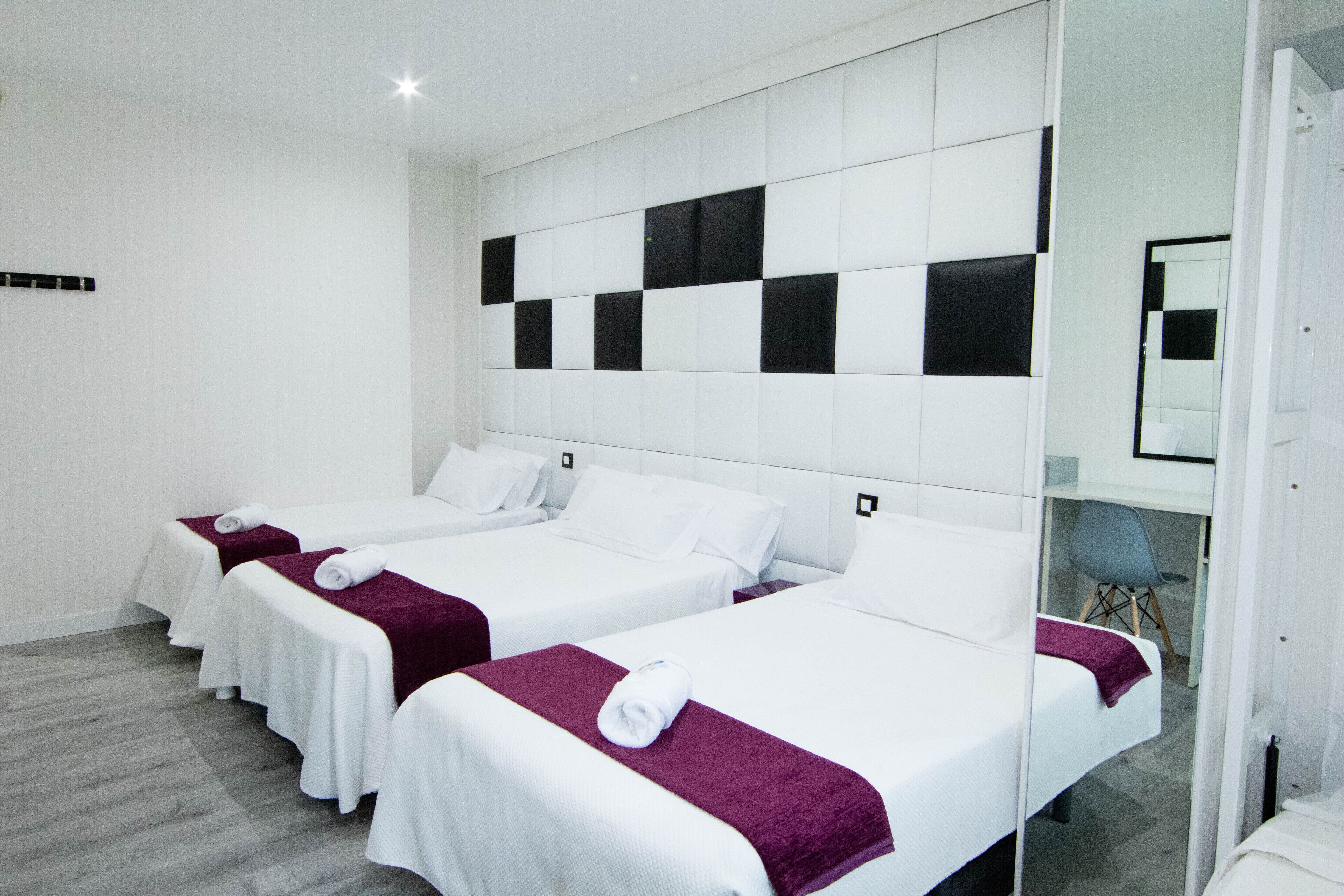 Habitación de hotel con tres camas, cada una con colcha blanca y manta morada. La habitación tiene un diseño moderno, con cabecera blanca y un espejo grande en la pared.