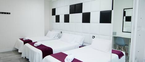 Une chambre d’hôtel avec trois lits, chacun recouvert d’une courtepointe blanche et d’un plaid violet. La chambre présente un design moderne, avec une tête de lit blanche et un grand miroir au mur.