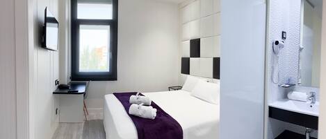 A modern hotel room with a bed, a desk, a large window, a bathroom with a mirror, and a towel rack.