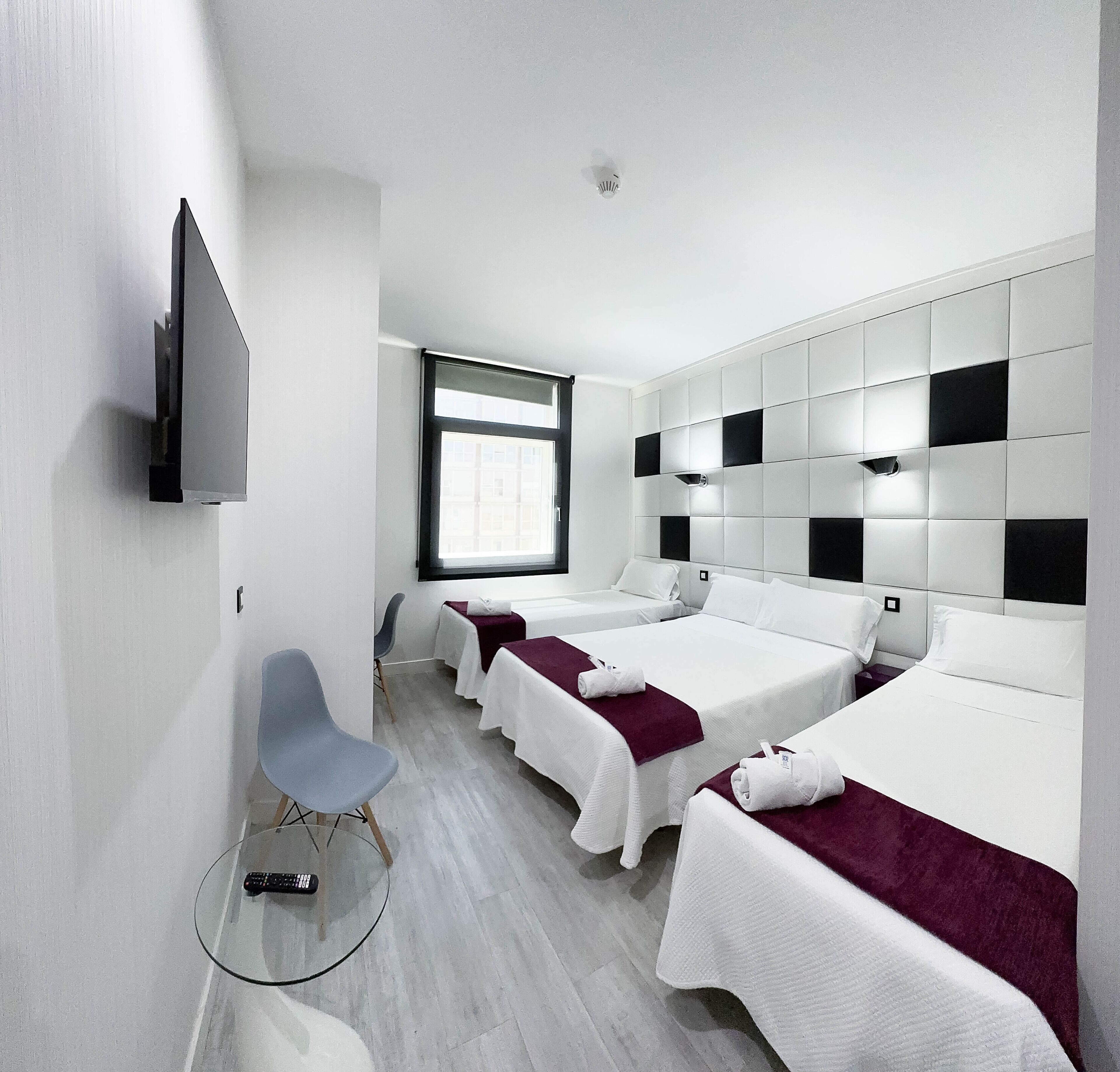 A modern hotel room with two single beds, a TV mounted on the wall, a window, and a glass table.