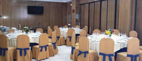 A banquet hall with round tables set for dining, each with chairs covered in beige fabric and blue sashes.