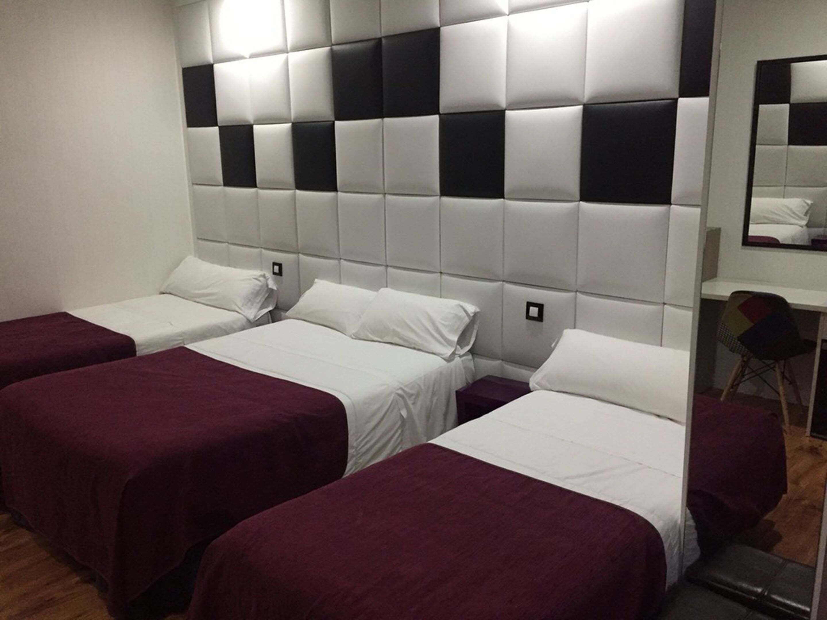 A hotel room with two beds, each with a white mattress and maroon bedspread. The headboard is white with black square accents.