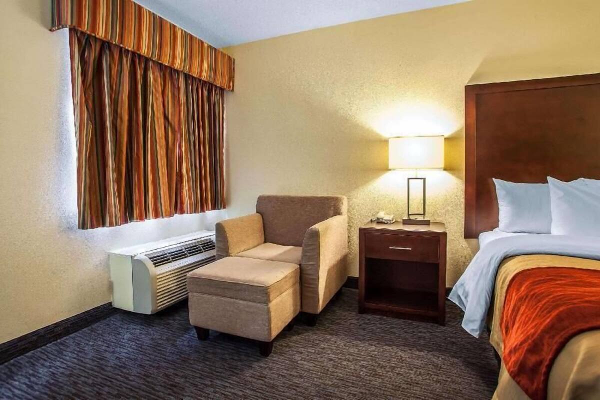 A hotel room with a bed, a chair, a nightstand, a lamp, and a window with curtains.