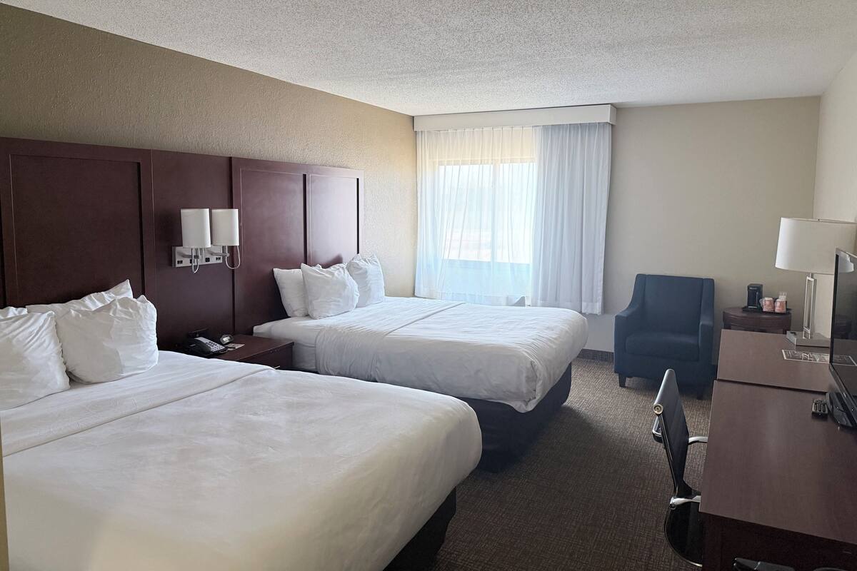 A hotel room with two beds, a desk, a chair, a lamp, and a window with curtains.
