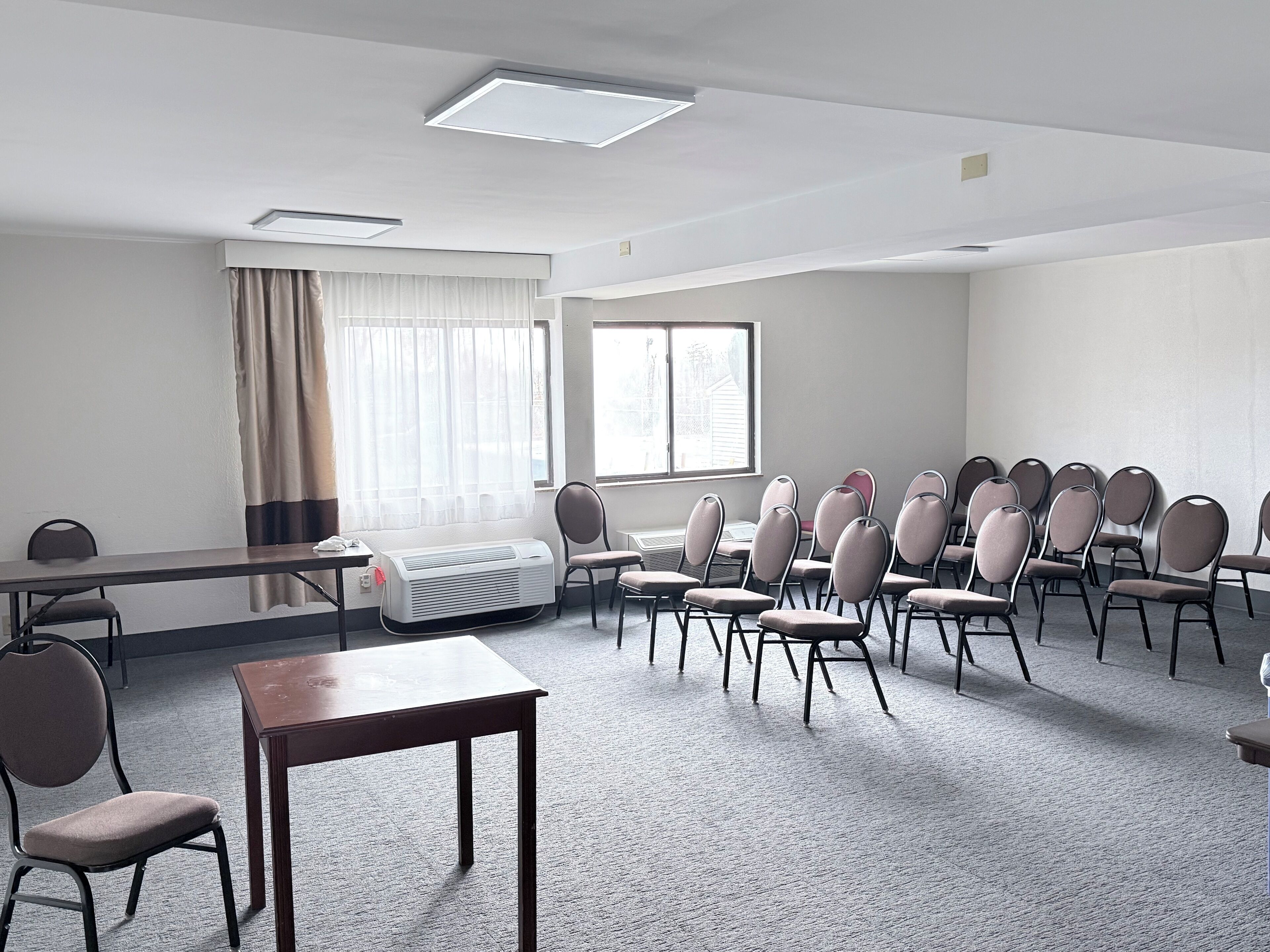Un salón de conferencias con una larga mesa, varias sillas, un ventanal con cortinas y una lámpara en el techo.