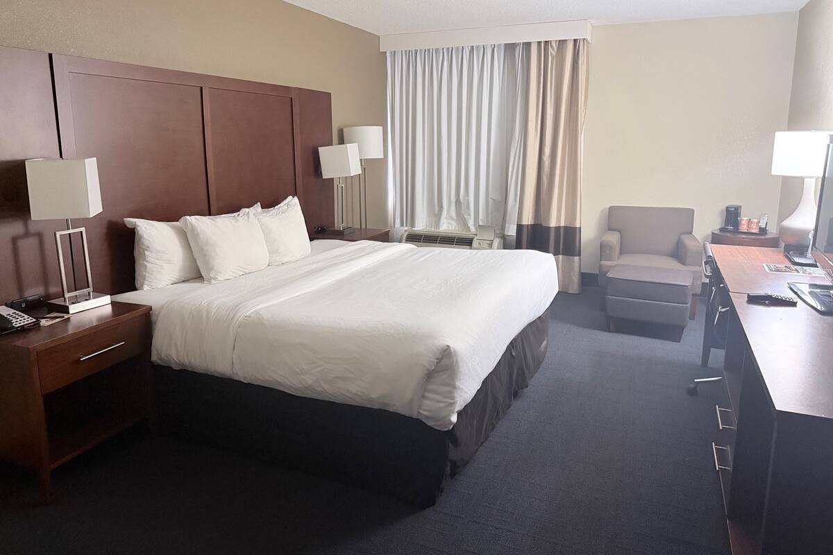 A hotel room with a large bed, a desk with a computer, a chair, a lamp, and a window with curtains.