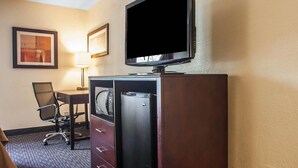 A hotel room with a flat-screen TV, a desk with a chair, a refrigerator, and a lamp.