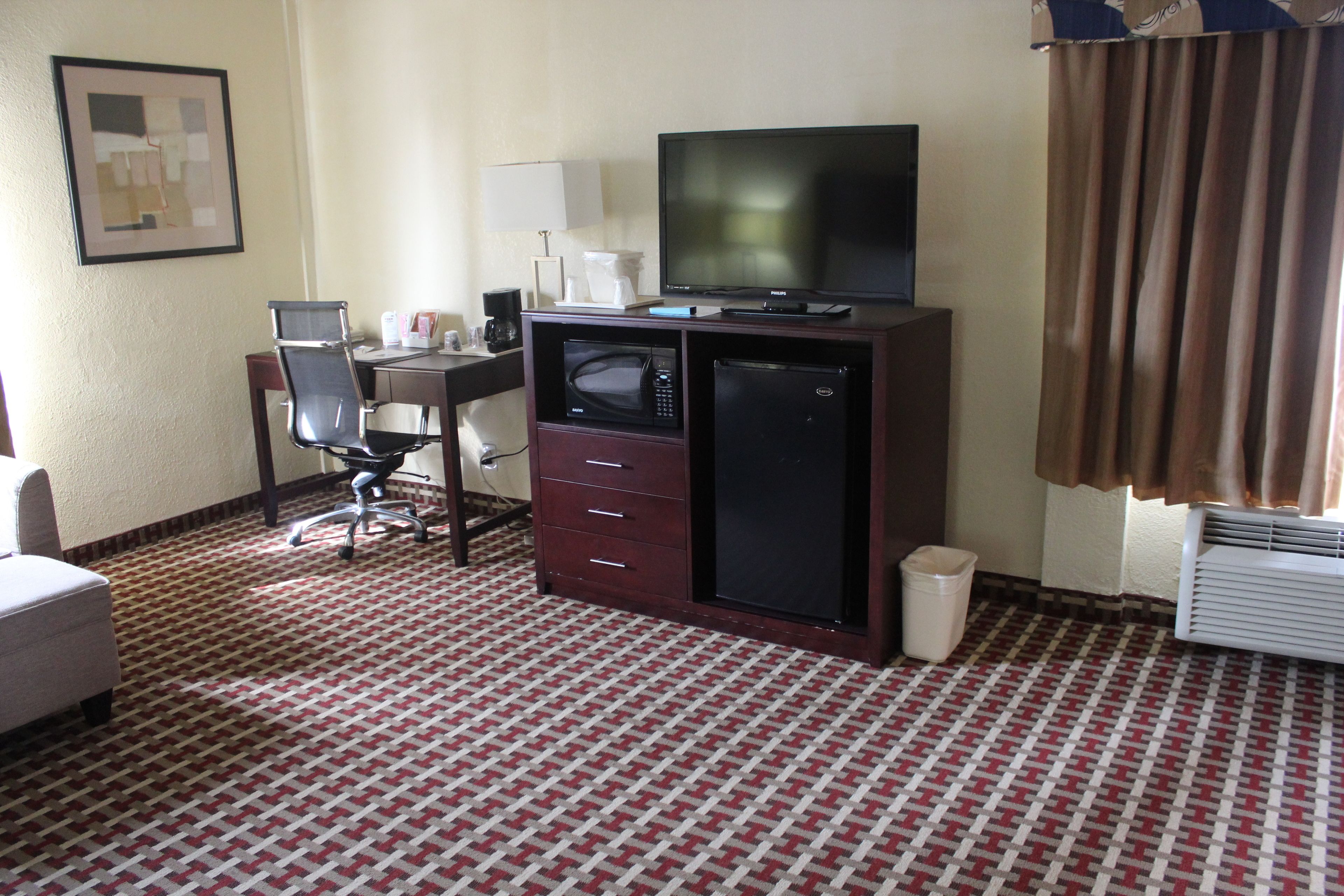 A hotel room with a desk, chair, television, microwave, and air conditioning unit.