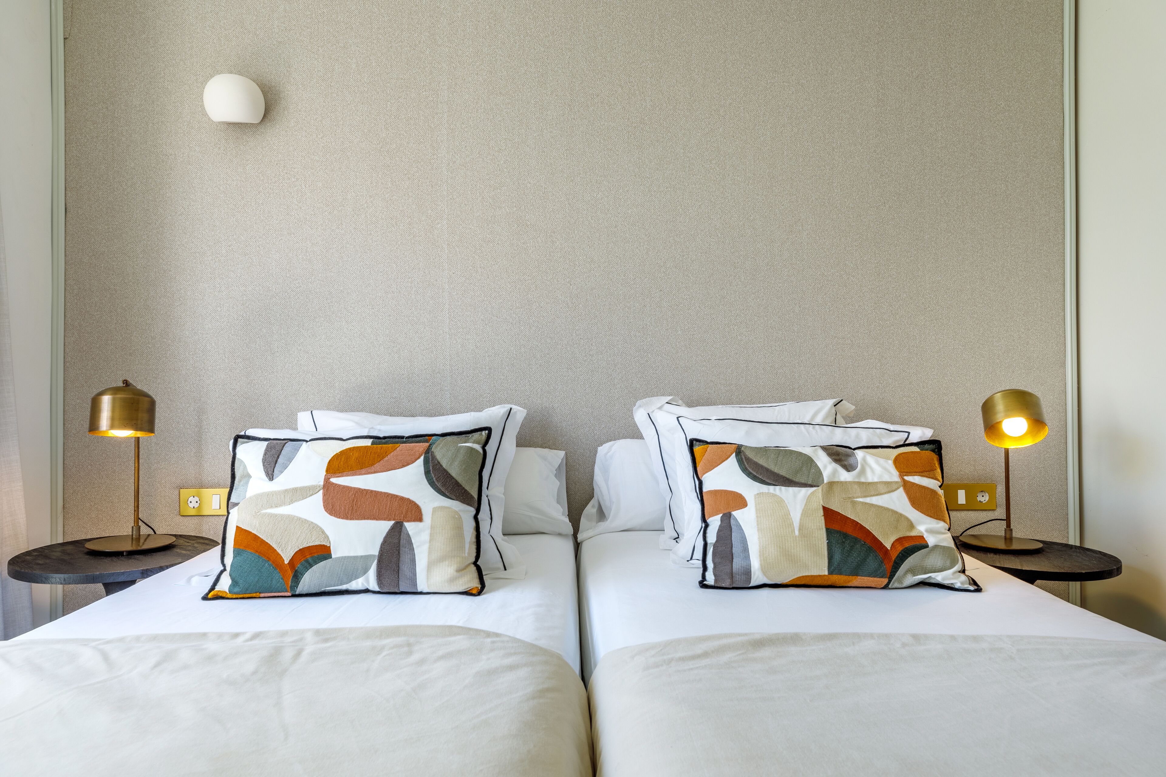 A hotel room with two beds, each with a patterned pillow, two bedside lamps, and a plain wall.