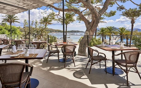 An outdoor dining area with tables and wicker chairs, overlooking a marina and a mountainous landscape.