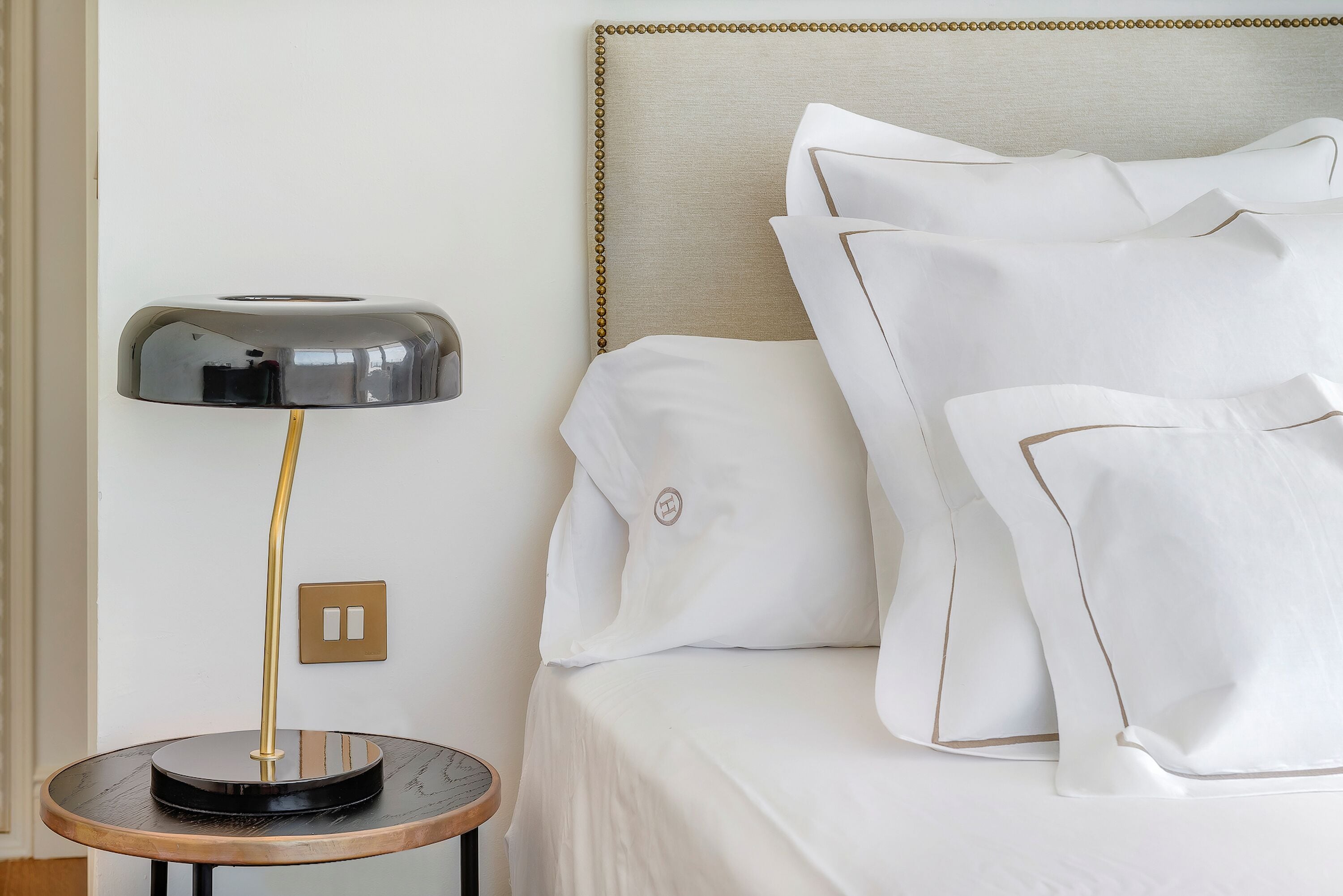 A neatly made bed with white linens and pillows, a round table with a lamp, and a wall-mounted light switch.