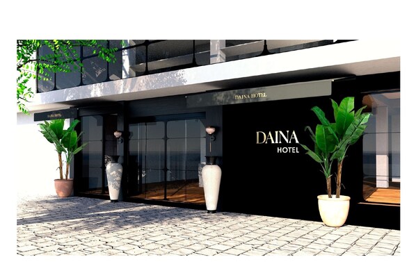 Exterior del hotel Daina, con un diseño moderno, que presenta grandes puertas de vidrio, plantas en macetas y un llamativo cartel con el nombre del hotel.