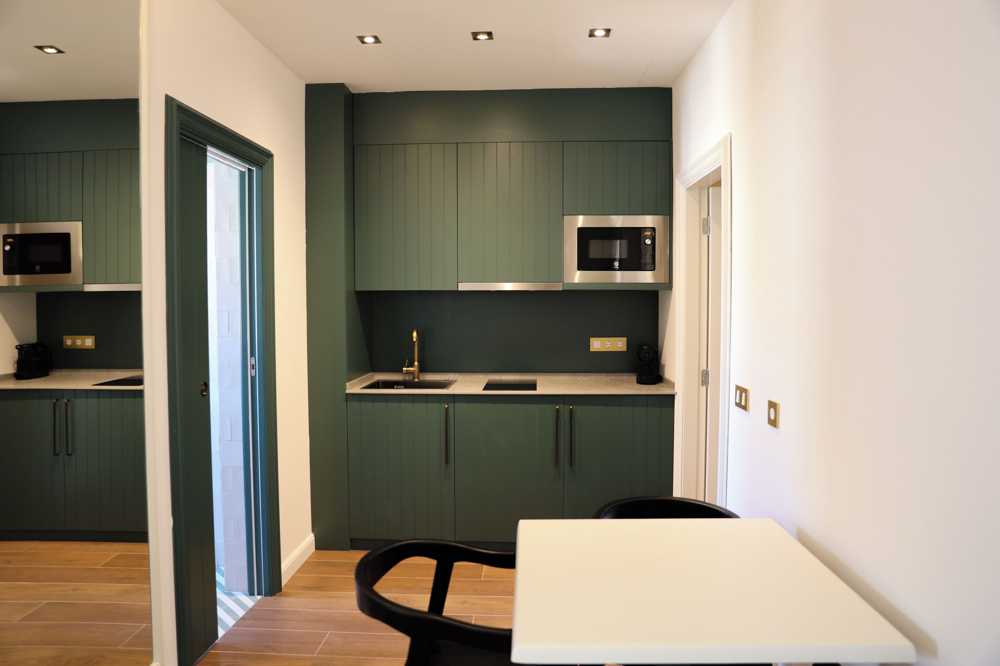 A compact kitchen with dark green cabinets, a white countertop, and a black chair.