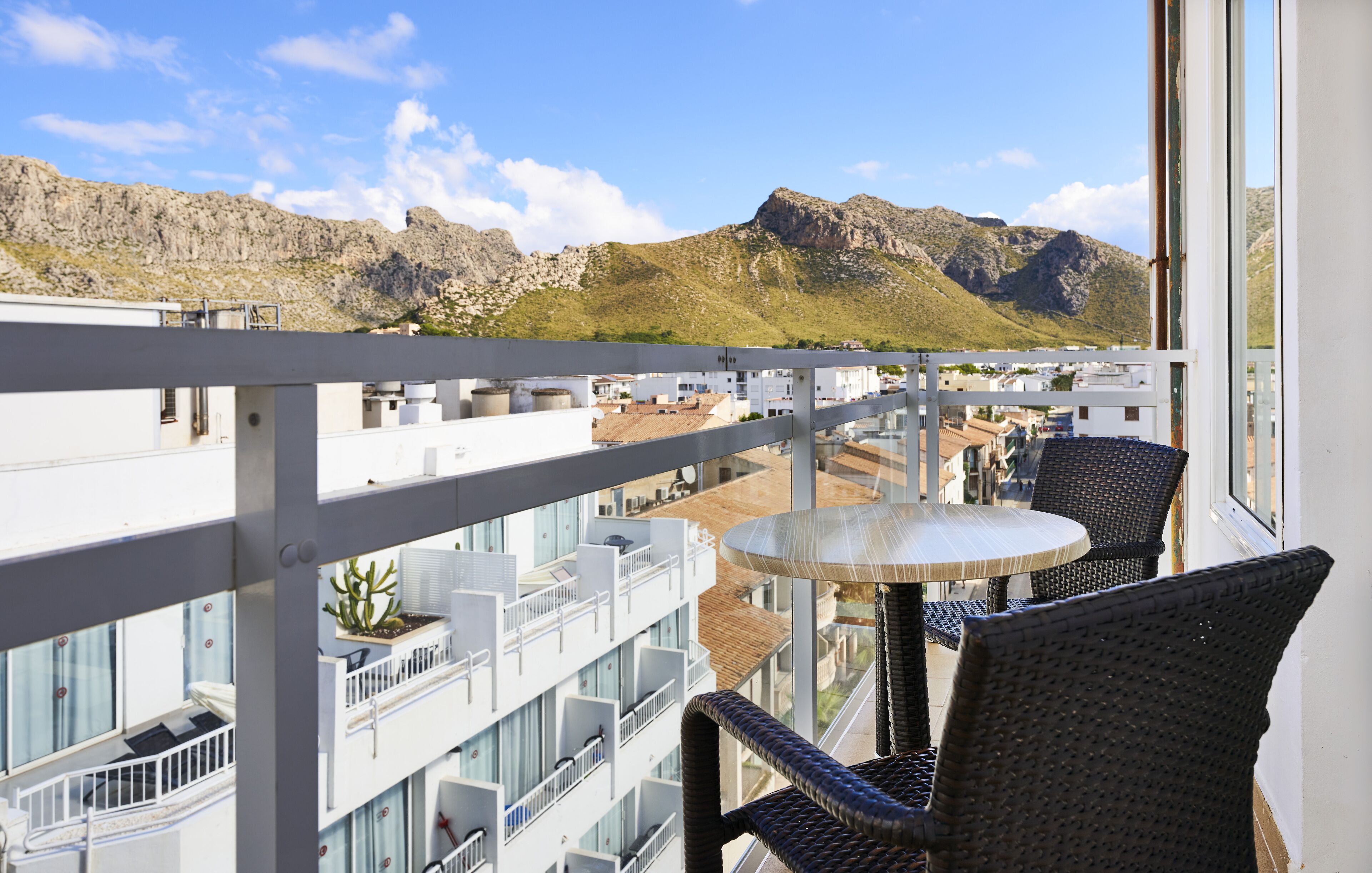 Ein Balkon mit Korbstühlen und einem kleinen Tisch, mit Blick auf eine gebirgige Landschaft und Gebäude im Hintergrund.