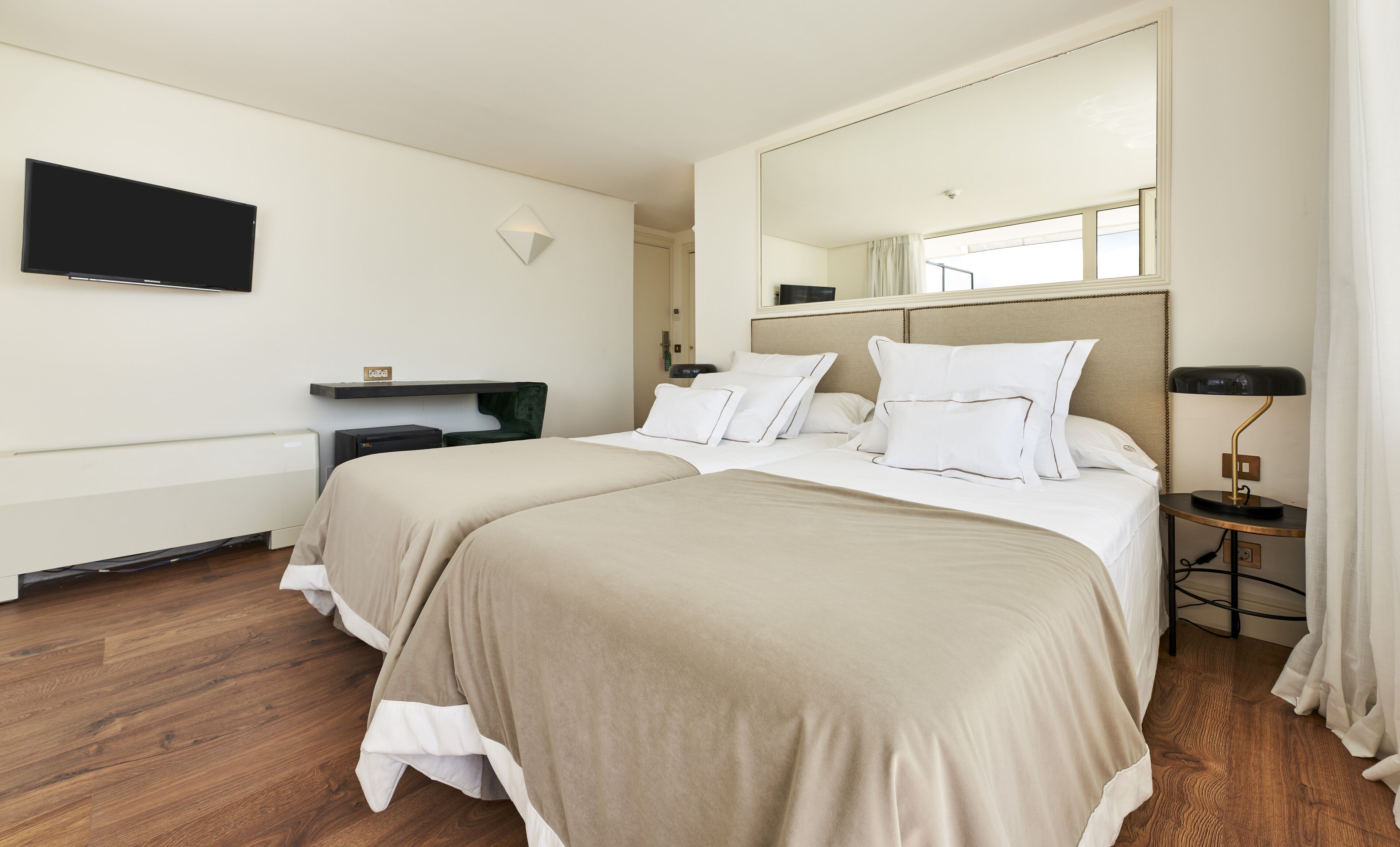 A hotel room with two beds, a flat-screen TV, a bedside table with a lamp, and a large mirror.
