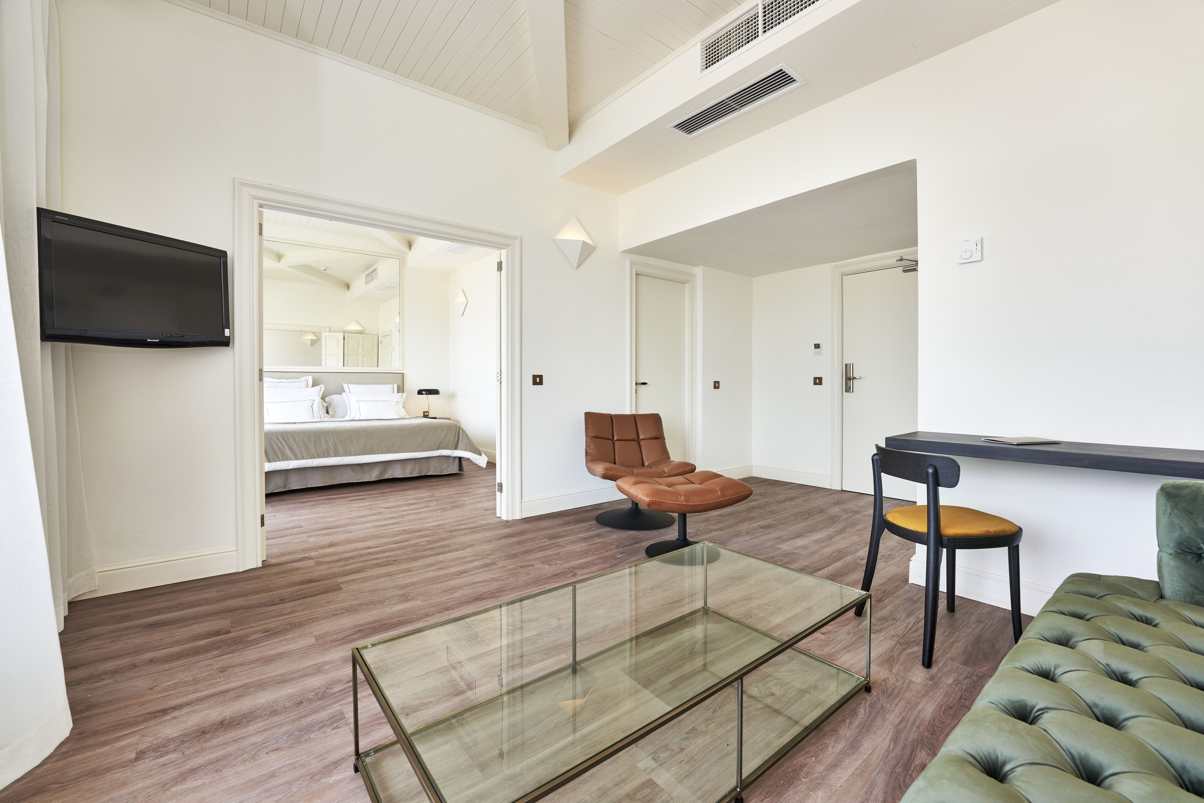 Une chambre d’hôtel moderne avec un lit, une table basse en verre, un fauteuil en cuir marron, un canapé vert et un téléviseur à écran plat fixé au mur.