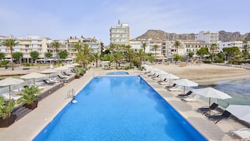 Una piscina rectangular con sillones de playa y sombrillas, rodeada de palmeras y edificios con balcones.