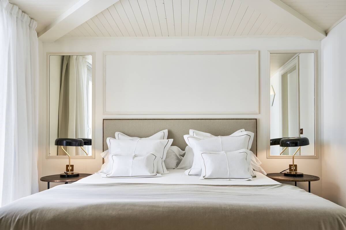 A neatly made bed with white linens and pillows, flanked by two bedside tables with lamps, in a room with a high ceiling and large mirrors on the walls.