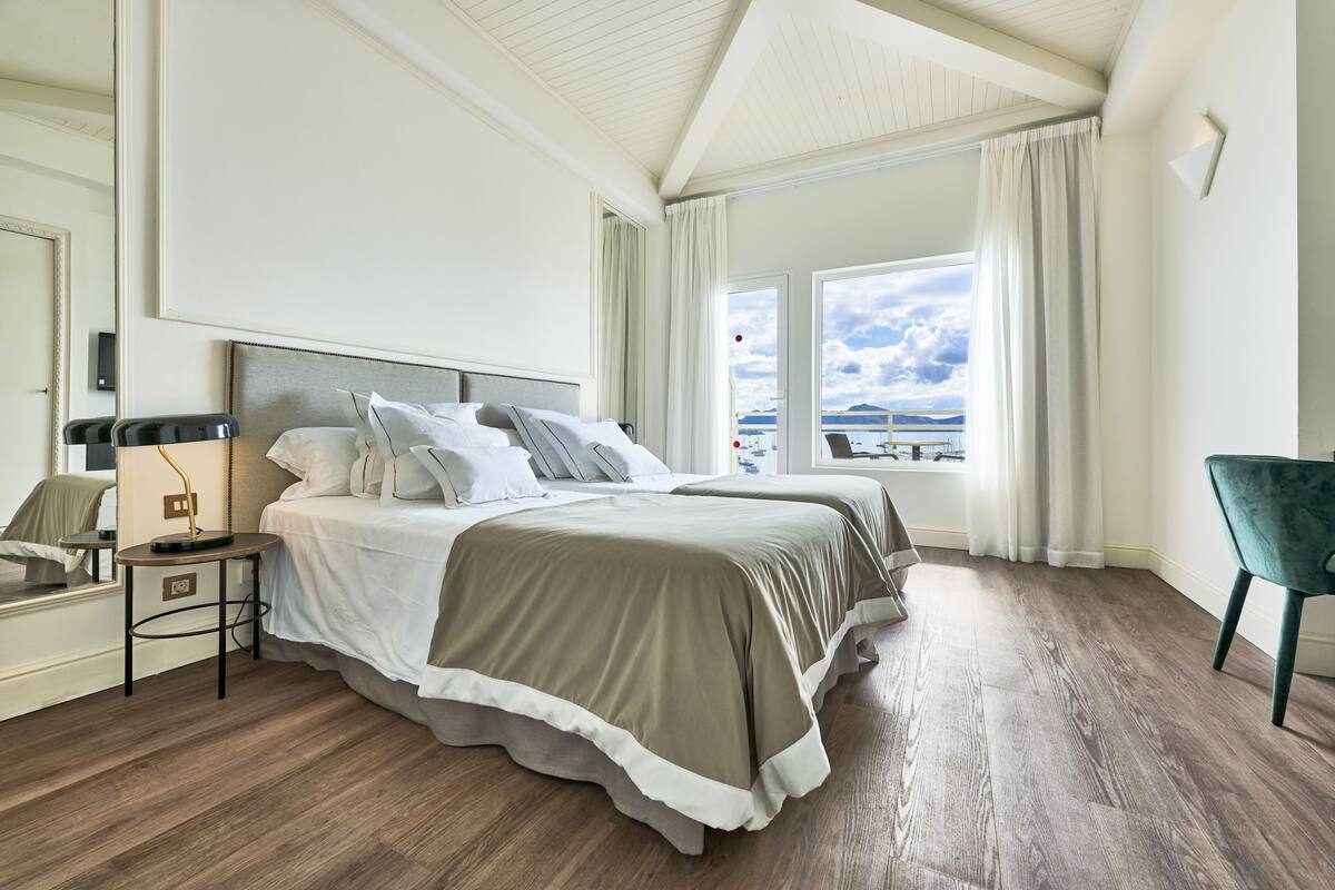 A bedroom with a large bed, a bedside table, a chair, and a view of the sea.